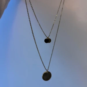 Silverfärgat halsband med dubbla kedjor - Elegant halsband med två tunna kedjor i silverfärg. Varje kedja har en rund, slät berlock som hänger i olika längder för en stilren look. Perfekt för att lägga till en subtil glans till din outfit.
