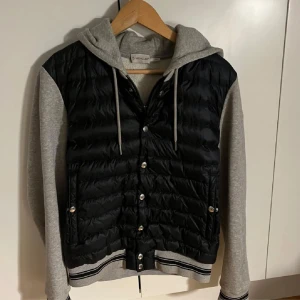 Svart och grå jacka med huva från Moncler - Säljer en snygg maglia cardigan från Moncler med svart quiltad framsida och grå ärmar och huva. Jackan har knappar och ribbade detaljer vid ärmslut och nederkant. Perfekt för våren/sommaren och skicket avgör du. Finns någon fläck på insidan samt liten på utsidan. Cond 8/10 därav priset. Säljes för att den inte kommer till användning!