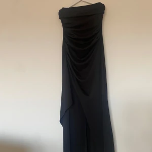 Svart axelbandslös långklänning - Elegant svart axelbandslös långklänning med draperad design framtill. Perfekt till en elegant kväll som bal.