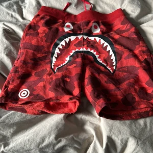 Röda camo shorts från A Bathing Ape - Snygga röda camo shorts från A Bathing Ape med ett coolt hajmotiv framtill. Shortsen har en elastisk midja med snörning och färgglada bokstäver på sidan. Perfekta för en avslappnad stil med en unik twist.