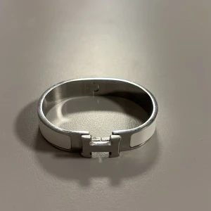 Hermés armband - Elegant armband i silverfärg med vit emaljdetalj och ett stilrent H-formad lås. Perfekt för att ge en sofistikerad touch till din outfit.