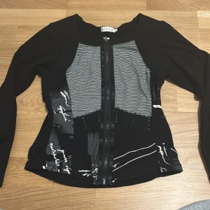 Svart mönstrad tröja från Micha - Snygg svart tröja från Micha med unikt mönster i vitt och grått framtill. Tröjan har en dragkedja och långa ärmar, perfekt för en stilren look. Passar bra till både jeans och kjol.