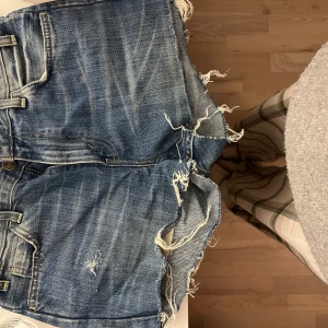 Blå lågmidjade jeansshorts från Levi's - Säljer ett par coola blå lågmidjade jeansshorts från Levi's med slitna detaljer och fransiga kanter. Shortsen har en klassisk femficksdesign och knappgylf. 