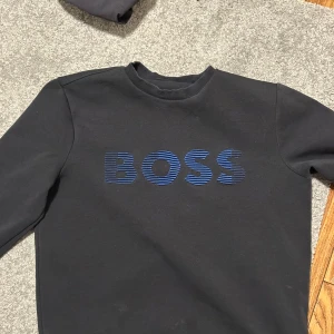 Svart tröja från BOSS - Säljer en stilren svart tröja från BOSS med blått logotryck på bröstet. Tröjan har en klassisk rund halsringning och långa ärmar. Perfekt för en avslappnad och trendig look.
