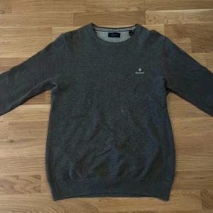 Grå tröja från GANT - Säljer en stilren grå tröja från GANT med rund halsringning. Tröjan har långa ärmar och ett diskret GANT-logotyp broderat på bröstet. Perfekt för en avslappnad och snygg look.