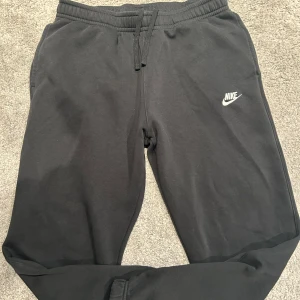 Svarta mjukisbyxor från Nike - Säljer ett par svarta mjukisbyxor från Nike med elastisk midja och snörning. Byxorna har en broderad Nike-logga på sidan och är perfekta för en avslappnad stil.