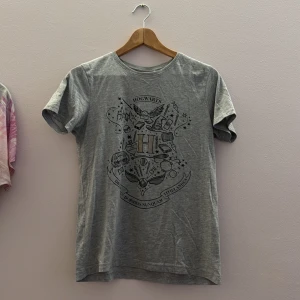 Grå t-shirt med Hogwarts-tryck - Säljer en grå t-shirt med ett detaljerat Hogwarts-tryck på framsidan. T-shirten har korta ärmar och är tillverkad i ett mjukt material. Perfekt för alla Harry Potter-fans!