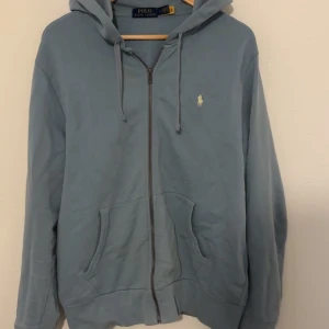 Blå hoodie från Polo Ralph Lauren - Säljer en stilren blå hoodie från Polo Ralph Lauren med dragkedja och huva. Tröjan har en broderad logotyp på bröstet och praktiska fickor framtill. Perfekt för en avslappnad look. Skick 9/10 helt ny.