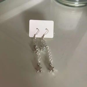 Silverfärgade stjärnörhängen - Säljer ett par snygga silverfärgade örhängen med hängande stjärnor. De har en kedjedesign som ger en cool och trendig look. Perfekta för att piffa upp vilken outfit som helst!