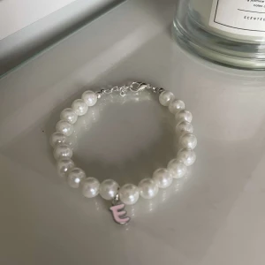 Pärlarmband med bokstavsberlock - Säljer ett superfint pärlarmband med vita pärlor och en söt rosa bokstavsberlock. Perfekt för att ge en personlig touch till din outfit. Armbandet har en enkel och elegant design som passar till alla tillfällen.