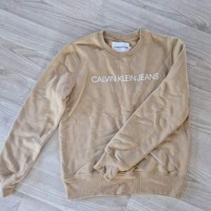 Säljer en snygg beige sweatshirt från Calvin Klein Jeans. Den är i storlek S och har en klassisk rund halsringning. Tröjan är långärmad och har ribbade muddar vid ärmslut och nederkant. Perfekt för en avslappnad och stilren look!