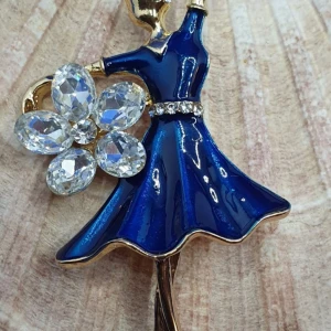 Brosch - Dansös i blå klänning - Den här broschen i form av en dansös i blå klänning är ca 7 × 3.7 cm. Under dansösens arm finns en stor blomma i kristall. 