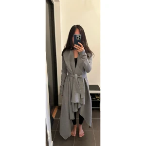 Grå lång kappa med bälte - Min älskade grå oregelbunden kappa från Norma Kamali - perfekt för höst och vår. Köpt för 3500 kr, är helt ny och oanvänd. Säljer pga jag är för kort för den. Storlek 32 (XXS) trots att jag brukar ha 34 (XS). Perfekt för dig som är lång och smal! 