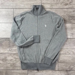 Grå kofta från Polo Ralph Lauren - Säljer en stilren och bekväm grå kofta från Ralph Lauren. Den har en dragkedja framtill och är långärmad med ribbade muddar. Perfekt för både höst och vår, och den är i nyskick. Den klassiska loggan är broderad på bröstet. Modellen är 178 o bär S
