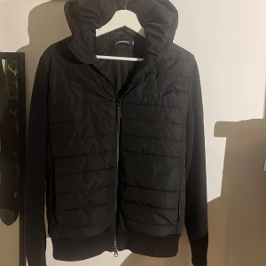 Svart jacka från J.Lindeberg - Snygg svart jacka från J.Lindeberg med quiltad design och dragkedja framtill. Jackan har en huva och ribbade muddar vid ärmslut och nederkant. Perfekt för kyligare dagar.nypris 3000kr skick 9/10.