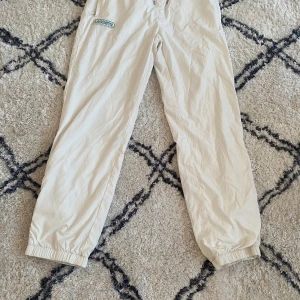 Vita byxor från Lacoste - Snygga beige byxor från Lacoste med elastisk midja och snörning. Byxorna har en avslappnad passform och är perfekta för en casual look. De har fickor fram och en bakficka med det klassiska Lacoste-märket.