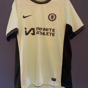 Chelsea fotbollströja - Chelsea fotbollströja från Nike med korta ärmar. Tröjan har Chelsea-loggan på bröstet och texten 'Infinite Athlete' tryckt på framsidan. Perfekt för alla Chelsea-fans!