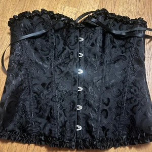 Svart korsett med broderat mönster - Elegant svart korsett med vackert broderat mönster och knäppning framtill. Den har en dekorativ snörning upptill och volangdetaljer längs kanterna. Perfekt för en stilfull look.