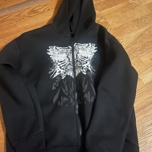 Svart hoodie med tryck - Snygg svart hoodie med dragkedja och ett unikt tryck i vitt på framsidan. Perfekt för en avslappnad stil med en cool edge. Den har en klassisk passform och en bekväm huva.