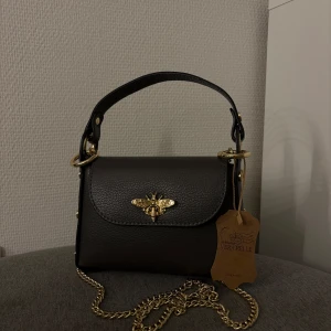 Svart handväska med gulddetaljer från Vera Pelle - Elegant svart handväska i skinn från Vera Pelle med en dekorativ guldfärgad bi-detalj på framsidan. Väskan har en kedja i guld och nitar på sidorna som ger en trendig look. Väskan är helt ny och gjord av italienskt läder. Väskan kan uppfattas som mörkare på bilderna💕