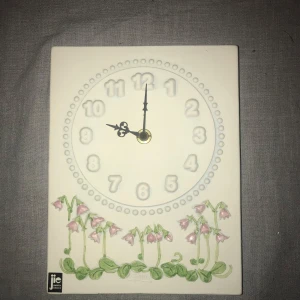 Jätte fin väggklocka från Jie Gantofta - Charmig väggklocka i keramik från Jie Gantofta med blommönster i rosa och grönt. Klockan har en rektangulär form och tydliga siffror i en cirkel. Perfekt för att ge en romantisk touch till ditt hem.