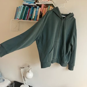Grön hoodie från Weekday - Säljer en stilren grön hoodie från Weekday. Den har en klassisk design med en stor ficka framtill och justerbar huva med dragsko.