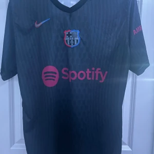 Svart FC Barcelona fotbollströja - Svart FC Barcelona tröja. Tröjan har klubbens emblem och Nike-logga i rött och blått. På baksidan finns namnet 'Lamine Yamal' och nummer 27. Väldigt skönt material och sitter bra om ärmarna. Vid snabba affärer kan sänka priset