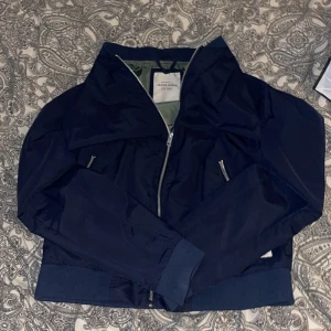 Mörkblå bomberjacka från Jack & Jones - Säljer en stilren mörkblå bomberjacka från Jack & Jones. Jackan har dragkedja framtill och två snedställda fickor med dragkedjor. Perfekt för en casual look.