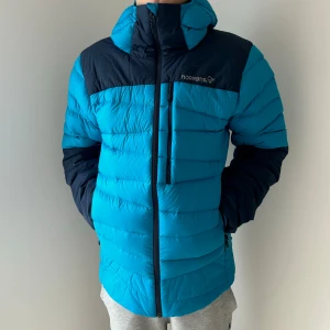 Blå dunjacka från Norrøna - Snygg blå dunjacka från Norrøna med svarta detaljer. Jackan har en hög krage och dragkedja framtill. Perfekt för kyliga dagar.