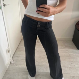 Svarta lågmidjade kostymbyxor  - Snygga svarta byxor med låg midja och två dekorativa knappar framtill. Midjemått 37 rakt över och innerbenslängden 77 cm