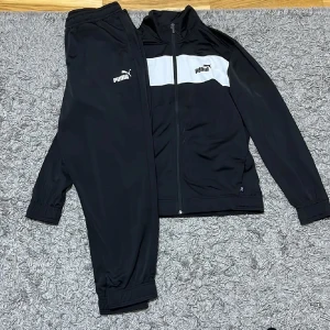 Svart tracksuit från Puma - Snygg svart tracksuit från Puma med vit panel och logga på bröstet. Jackan har dragkedja framtill och långa ärmar. Perfekt för sport och träning.