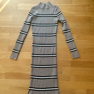 Randig ribbad klänning från New Look - Snygg ribbad klänning från New Look i beige med svarta och vita ränder. Den har en hög krage och långa ärmar, perfekt för en stilren look. Klänningen är figurnära och sträcker sig till knälängd.