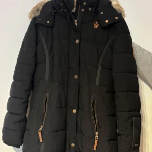 Svart dunjacka med pälsdetaljer från Marikoo - Snygg svart dunjacka från Marikoo med avtagbar huva och pälsdetaljer. Jackan har dragkedja och knappar framtill samt praktiska fickor med dragkedjor. Perfekt för kalla vinterdagar.