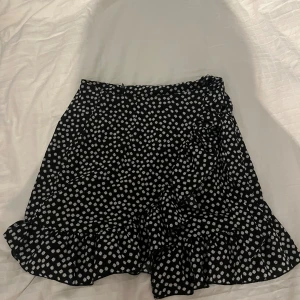 Svart blommig kjol från Shein - Snygg svart kjol från Shein med vita blommor och volangdetaljer. Kjolen har en dragkedja i sidan och är perfekt för en trendig look.