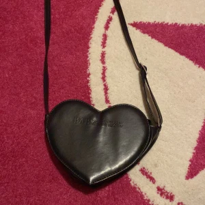 Svart väska från Baby, The Stars Shine Bright - Snygg svart axelväska i hjärtform från Baby, The Stars Shine Bright. Väskan har en justerbar rem och stängs med dragkedja. Perfekt för att ge en söt touch till din outfit. Tillverknings år 2013