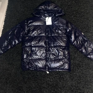Marinblå pufferjacka från Moncler - Snygg svart pufferjacka från Moncler med glansig finish och huva. Jackan har en dragkedja framtill och en praktisk ficka med Moncler-logga på ärmen. Perfekt för kyliga dagar.                         PRIS KAN DISKUTERAS  