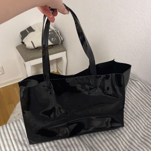 Svart glansig axelväska - Snygg svart axelväska i glansigt material. Perfekt för att bära med sig det nödvändigaste med stil. Väskan har två långa handtag som gör den enkel att bära över axeln. den är andvänd men funkar som ny med lite skorpor 