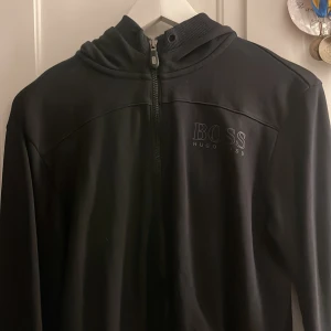 Svart tröja från Hugo Boss - Snygg svart tröjan från Hugo Boss med dragkedja och huva. Perfekt för en stilren look. Tröjan har långa ärmar och praktiska fickor framtill.