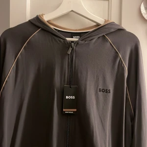 Grå hoodie från Boss - Snygg grå hoodie från Boss med dragkedja och huva. Den har diskreta kontrastlinjer längs ärmarna och en broderad logga på bröstet. Perfekt för en avslappnad stil.