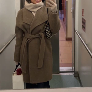 Beige kappa med bälte - Snygg och stilren beige kappa med knytbälte i midjan. Perfekt för kyliga dagar med sin knä långa design och breda krage. Ett måste för garderoben! Väldigt bra kvalité nopprar inte. 