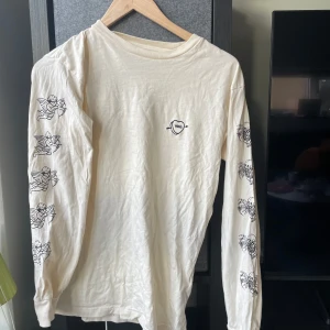 Vit långärmad tröja från Vans med skelettmönster - Säljer en cool vit långärmad tröja från Vans med unikt skelettmönster längs ärmarna. Perfekt för dig som gillar en edgy stil. Tröjan har en liten Vans-logga på bröstet i form av ett hjärta med pil.