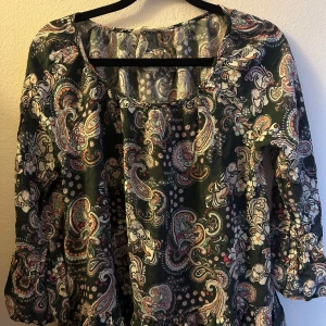 Mönstrad blus med trekvartsärm - Säljer en färgglad blus med paisleymönster i grönt, vitt och rött. Blusen har en rund halsringning och trekvartsärmar, perfekt för en bohemisk stil.