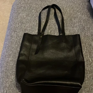 Svart axelväska i skinn - Snygg och rymlig svart axelväska i skinn med två handtag. Perfekt för att bära med sig allt man behöver under dagen. Väskan har en stilren design och stängs med en tryckknapp.