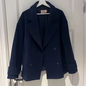 Mörkblå kappa från Nelly - Snygg mörkblå kappa från Nelly med dubbelknäppning och breda slag. Perfekt för kyligare dagar med sin stilrena design och långa ärmar. Defekt inuti kappan, söm som spruckit men går självklart att laga 
