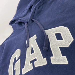 Mörkblå hoodie från GAP - Säljer en snygg mörkblå hoodie från GAP med stor logga framtill. Tröjan har en klassisk känguruficka och justerbar huva med dragsko. Perfekt för en avslappnad stil. Nyskick i princip. Inga tecken på användning sitter som storlek M
