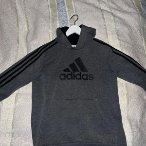 Grå hoodie från Adidas - Säljer en grå hoodie från Adidas med svart logga och de klassiska tre ränderna längs ärmarna. Tröjan har blivit för liten och behöver ränsa garderoben. Prister kan självklart diskuteras 