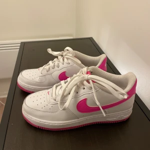 Nike Air Force med rosa detaljer - Snygga vita Nike Air Force med rosa detaljer och sula. Skorna har klassisk snörning och är perfekta för en sportig look. De har en ikonisk design med en färgglad twist.
