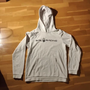 Vit hoodie från Sail Racing - Säljer en vit hoodie från Sail Racing med svart logotyp på bröstet. Tröjan har en klassisk känguruficka och en bekväm huva. Perfekt för en avslappnad stil. 