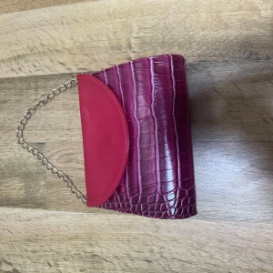 Röd axelväska med kedja - Snygg röd axelväska med krokodilpräglat mönster och kedjehandtag. Perfekt för att ge en färgklick till din outfit. Väskan har en stilren design med en elegant kedja som axelrem. Priset går att diskuteras 