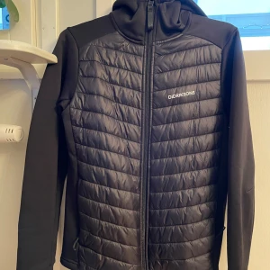 Svart jacka från Didriksons - Snygg svart cardigan från Didriksons med quiltad framsida och dragkedja. Jackan har en huva och långa ärmar, perfekt för kyligare dagar. Den är stilren och funktionell med en modern design.